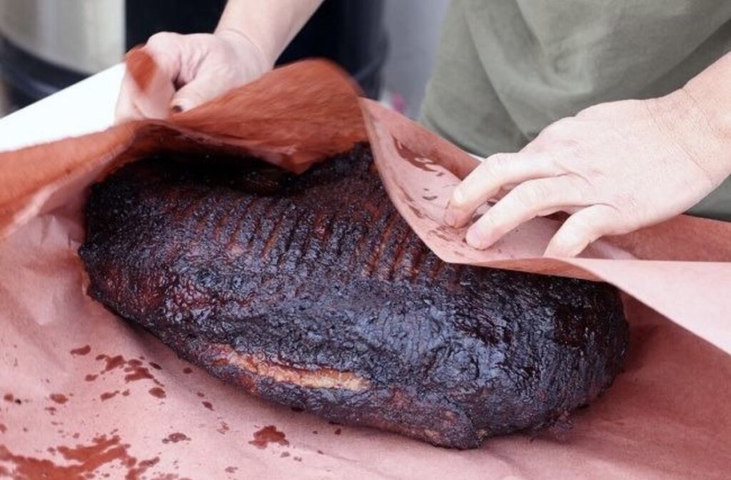 A person preparing a smoked brisket by peeling the bark.